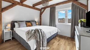 Ein Schlafzimmer mit einem großen Bett, Holzbalken, einem Fenster mit Vorhängen, einem Nachttisch mit Lampe und Blick ins Freie.