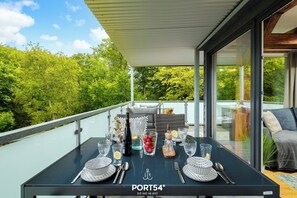 Une table à manger dressée avec de la verrerie, des assiettes et des couverts, sur un balcon offrant une vue sur les arbres et un intérieur moderne.