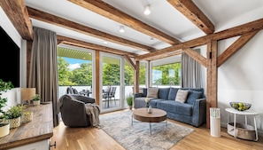 Una sala de estar moderna con vigas de madera, un sofá, una mesa de centro y vistas amplias a través de grandes ventanales.