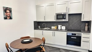 A modern kitchen with white cabinets, a microwave, a coffee maker, and a dishwasher. There is a round wooden table with chairs and a painting on the wall. - Captivating 2-bed Apartment in Brighton (Brighton)