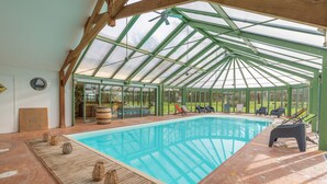 Una piscina techada con techo de vidrio, terraza de madera y sillones de descanso.