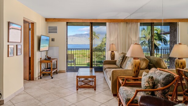 A spacious living room with a large sliding glass door leading to a balcony with a view of the ocean and palm trees. There is a sofa, two armchairs, a coffee table, and a TV on a stand.