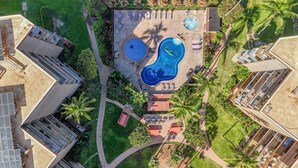 An aerial view of a resort with two swimming pools, a sun deck, and surrounding greenery.