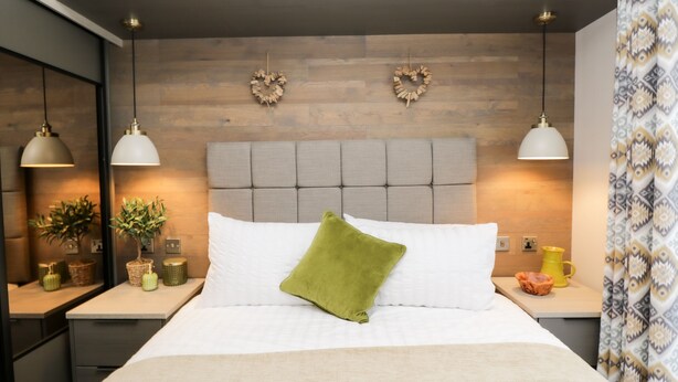 A modern bedroom with a tufted headboard, two pendant lights, a green pillow, and a bedside table with a plant and decorative items.