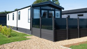 A modern mobile home with a large glass window, a dark exterior, and a gravel driveway.