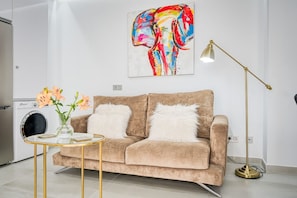A living room with a beige sofa, two white fluffy pillows, a glass coffee table, a vase with flowers, a washing machine, a colorful abstract painting, and a floor lamp. - Sun&beach en Las Canteras - 2 Bedroom Apartment (Las Palmas de Gran Canaria)