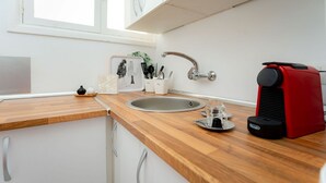 Una cocina con encimera de madera, pileta de acero inoxidable, cafetera roja y ventana con persianas.