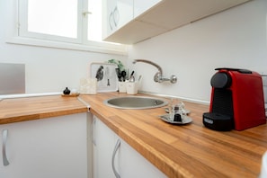 Una cocina con encimera de madera, pileta de acero inoxidable, cafetera roja y ventana con persianas.