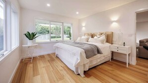 Een slaapkamer met een groot raam, een nachtkastje, een bed met witte beddengoed en een houten vloer.