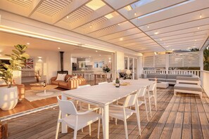 An outdoor patio with a wooden deck, white furniture, and a view of the interior.