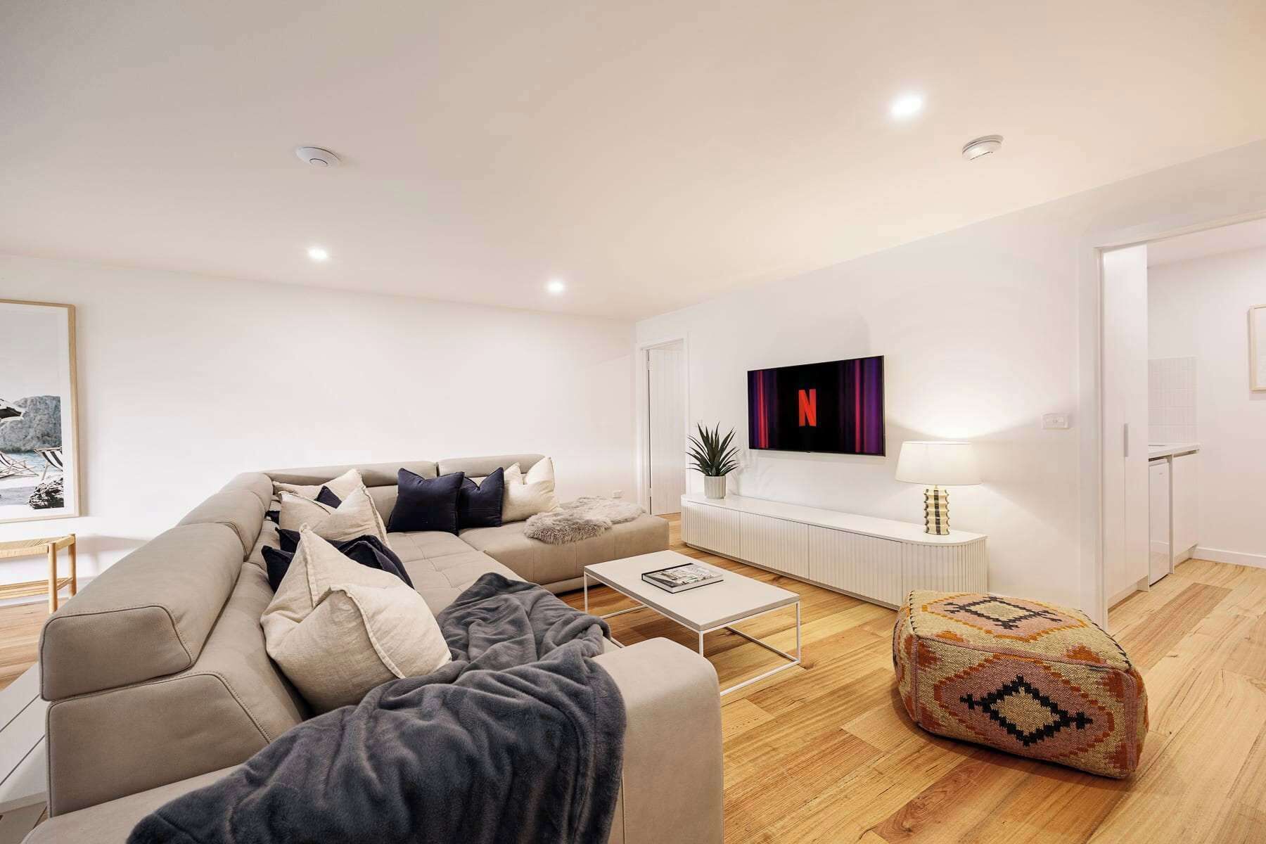 Un salon moderne avec un canapé, une table basse et un pouf. Un téléviseur est fixé au mur, ainsi qu’une lampe et une plante en pot.
