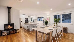 Een moderne keuken met een houtkachel, witte kasten en een eethoek met een houten tafel en witte stoelen.