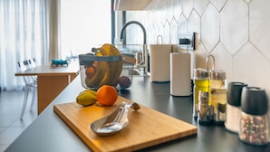 Une cuisine moderne avec un plan de travail en bois, un panier de fruits, une planche à découper, un couteau, un évier et une fenêtre avec des rideaux.