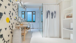 Une salle de bain moderne avec un mur orné d’un motif géométrique, un miroir, un rideau de douche à motifs de plumes et une vue sur l’extérieur par la fenêtre.
