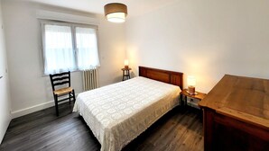 Quarto com cama de madeira, uma cadeira, janela com cortinas e mesa de cabeceira com abajur.