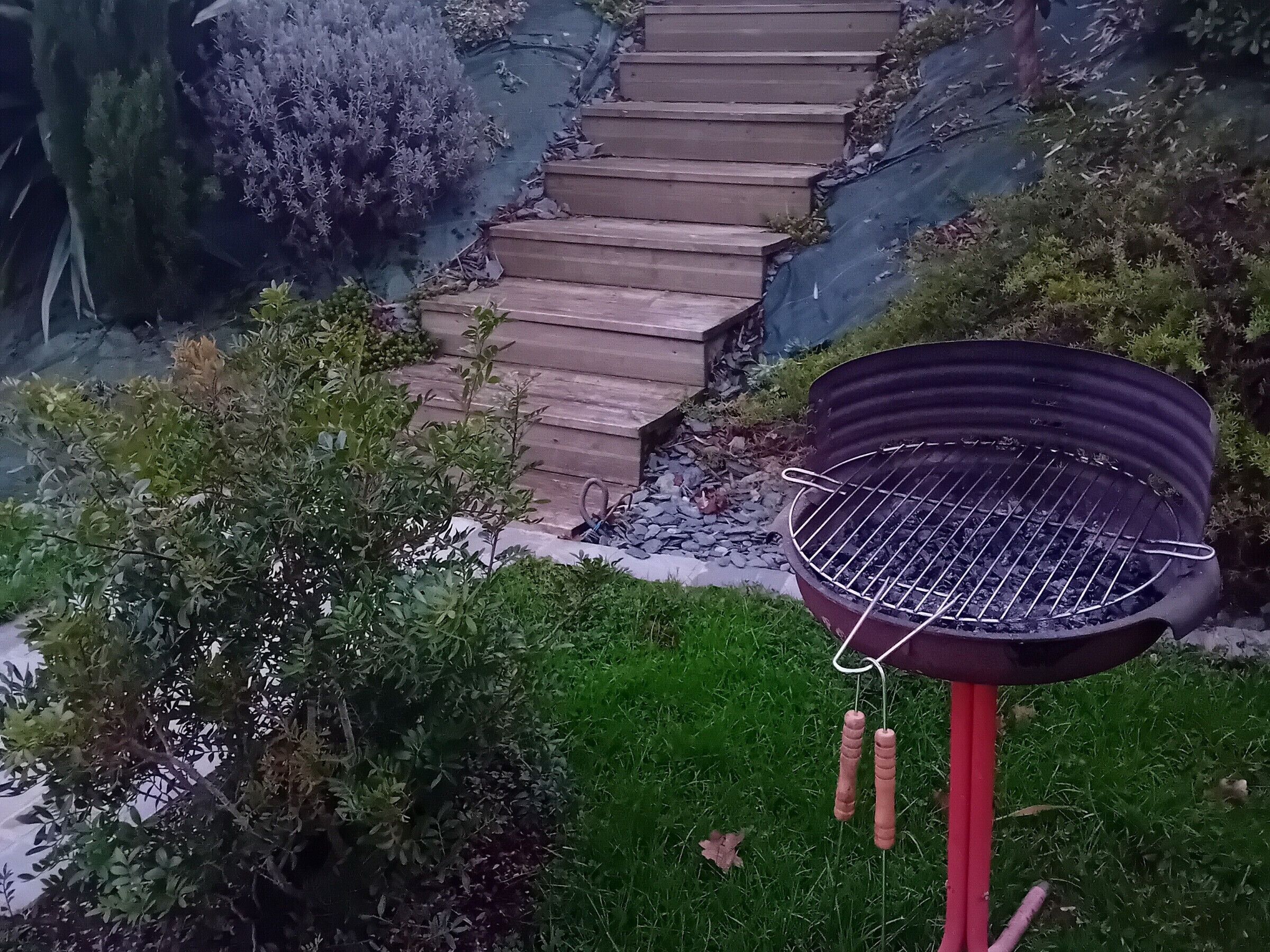 A barbecue grill with a metal grate and two handles, positioned on a grassy area with a wooden staircase in the background.