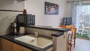 A modern kitchen with a black countertop, a built-in microwave, a coffee maker, and a sink. There is a painting of colorful parrots on the wall and a dining area with a table and chairs. - Beachside 2-BR Apartment for 5 with Private Parking & Terrace in Marseillan (Marseillan-Plage)