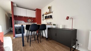 A compact kitchen with a dining area, featuring a built-in oven, black bar stools, and a small cabinet with a lamp. - Cozy Studio with Balcony, Sleeps 3, Near Slopes and Amenities (La Salle-les-Alpes)