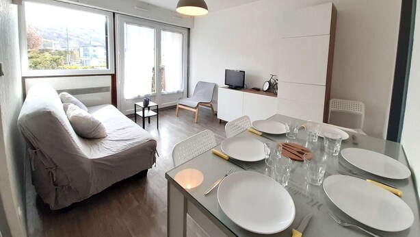 Un salon moderne et bien éclairé, avec un canapé, une table à manger pour quatre personnes et un téléviseur posé sur un meuble.