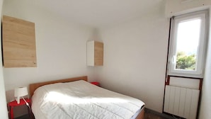 Une chambre à coucher avec un lit, une table de chevet, un tabouret rouge, une fenêtre et un radiateur.