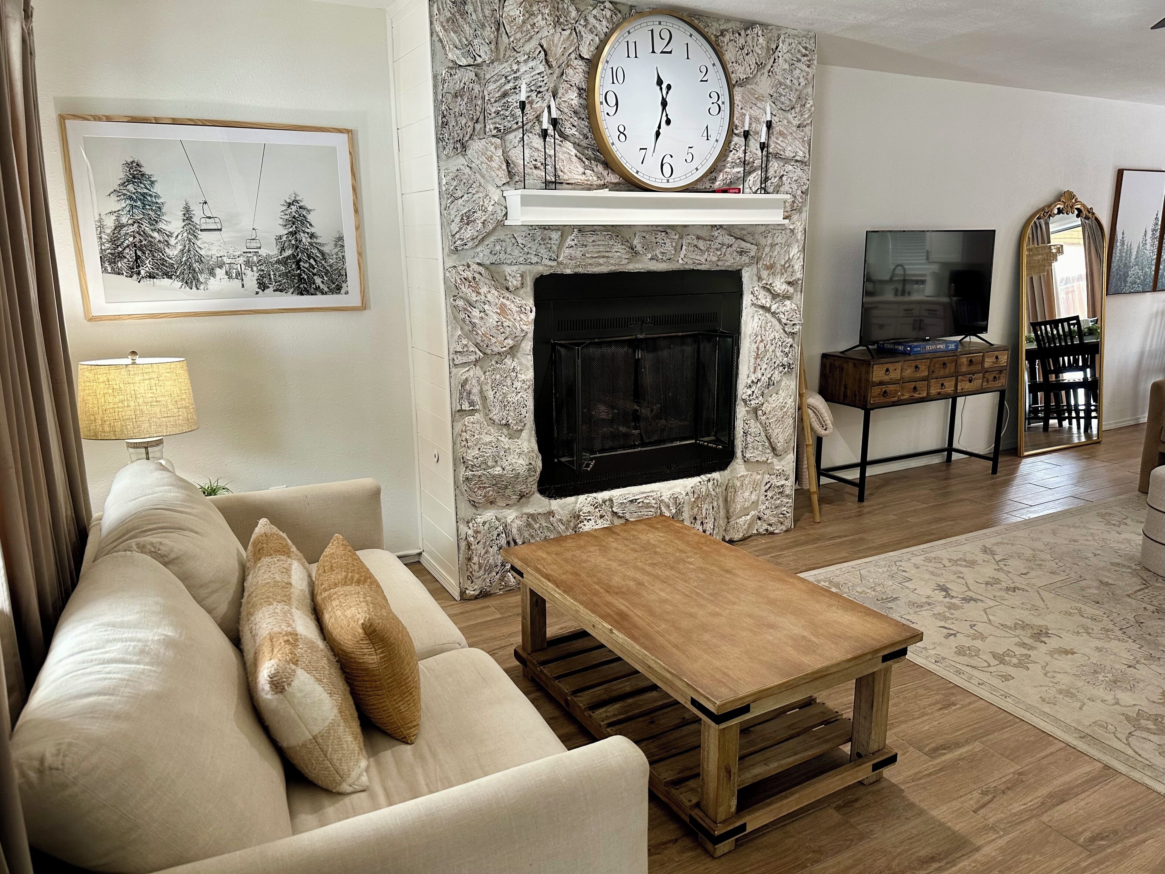 A living room with a stone fireplace, a sofa with cushions, a coffee table, a TV, and a mirror.