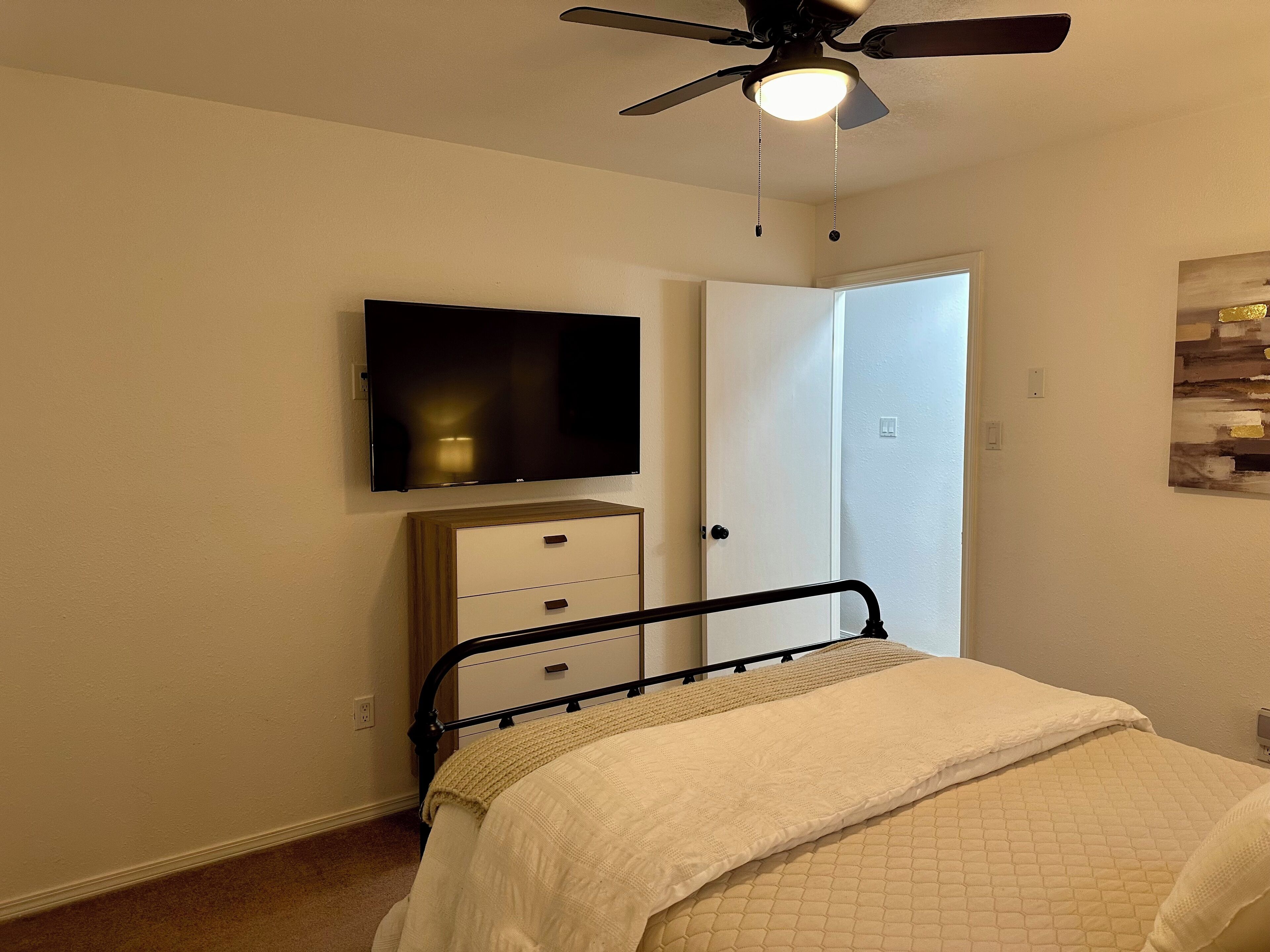 A bedroom with a bed, a dresser, a television mounted on the wall, and a ceiling fan.