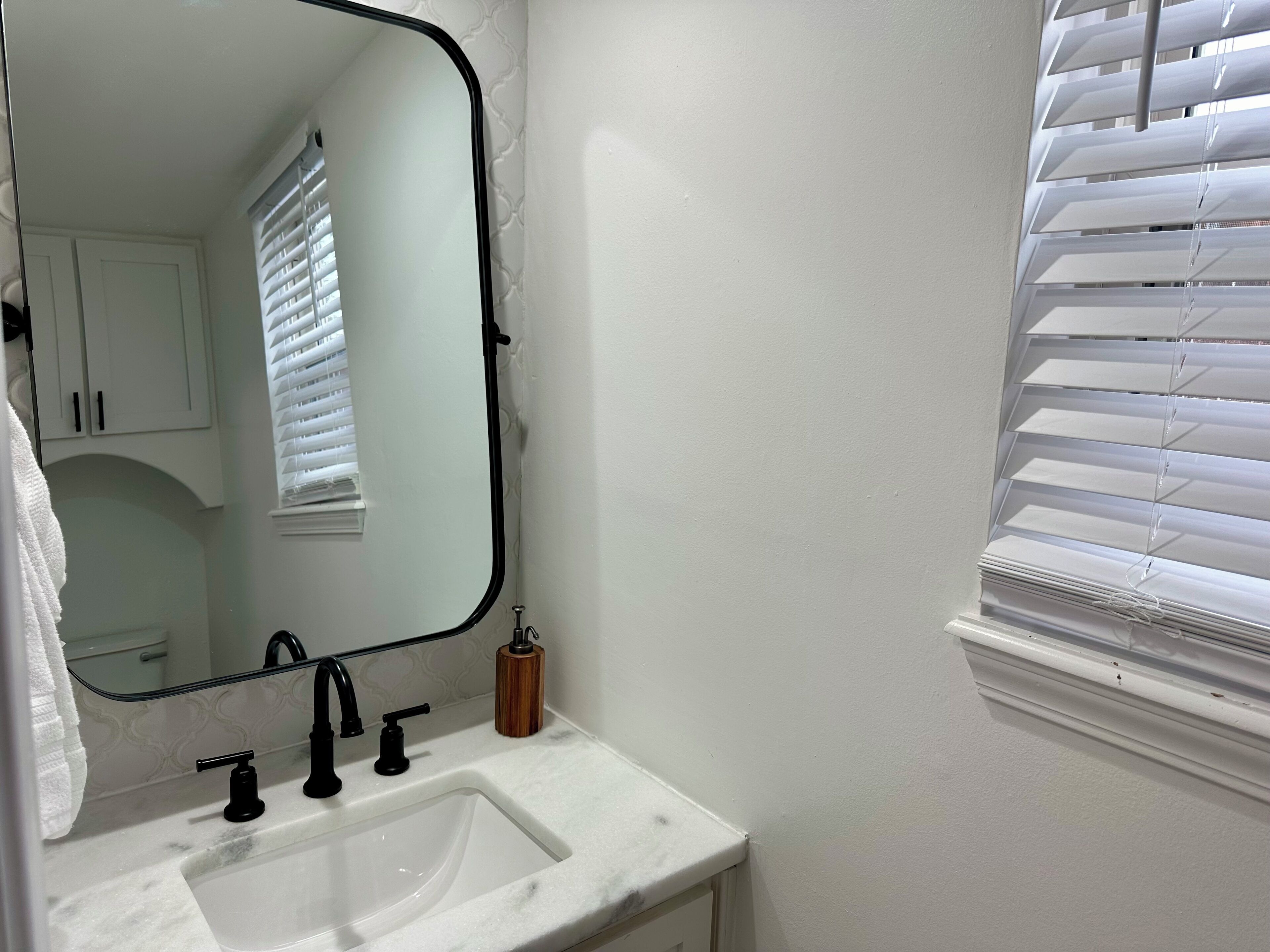 Een badkamer met een witte wastafel, zwarte kraanwerk, een spiegel met een zwarte lijst, een houten zeepdispenser en witte luxaflex.