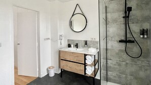 A modern bathroom with a glass shower enclosure, a round mirror, and a wooden vanity with towels. - Chambre Quadruple Avec Salle de Bains Privative à 10 Minutes de Disneyland Paris (Couilly-Pont-aux-Dames)