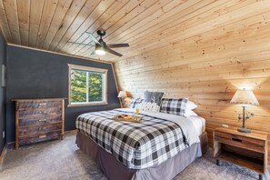 A bedroom with a bed, wooden ceiling, a dresser, a nightstand with a lamp, and a window with a view of trees. - Mountain Lake Lodge | Pet-Friendly, Bunkhouse + Lake Views | Near Yellowstone (Island Park)