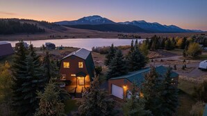 Eine Hütte mit großem Fenster, einer Veranda und einer Garage, umgeben von Bäumen und einem See vor einer Bergkulisse.