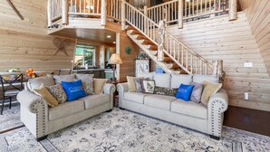 Ein geräumiger Wohnbereich mit hoher Decke und Holzwänden, ausgestattet mit zwei Sofas und dekorativen Kissen, sowie einer Treppe, die zu einer oberen Ebene führt.