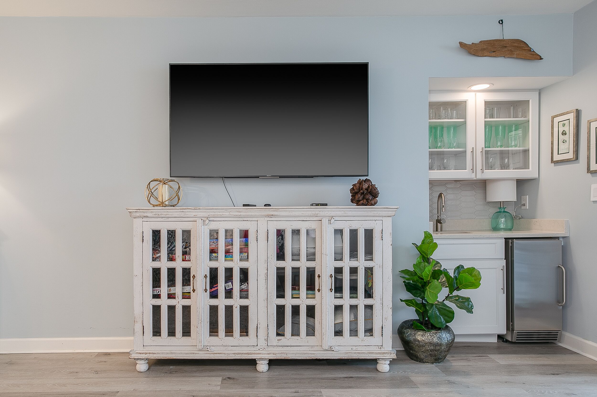 A living room with a flat-screen TV mounted on the wall, a white cabinet with glass doors, a small refrigerator, and a potted plant.