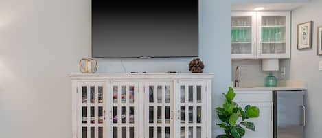 A living room with a flat-screen TV mounted on the wall, a white cabinet with glass doors, a small refrigerator, and a potted plant.