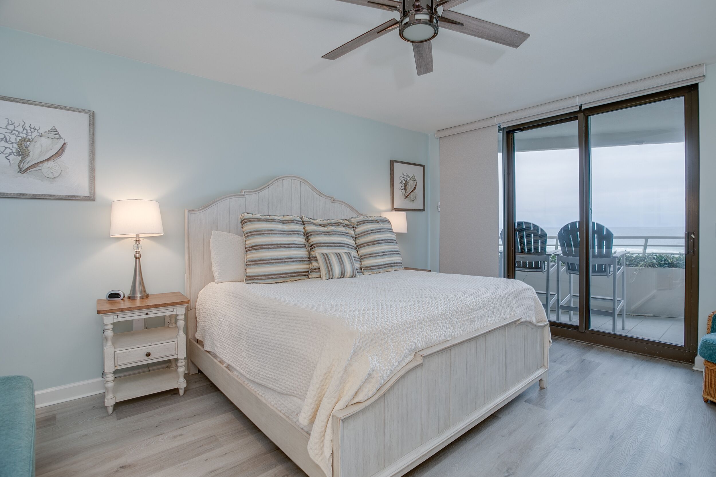 A bedroom with a bed, a nightstand, a ceiling fan, and a view of the sea through a sliding glass door.