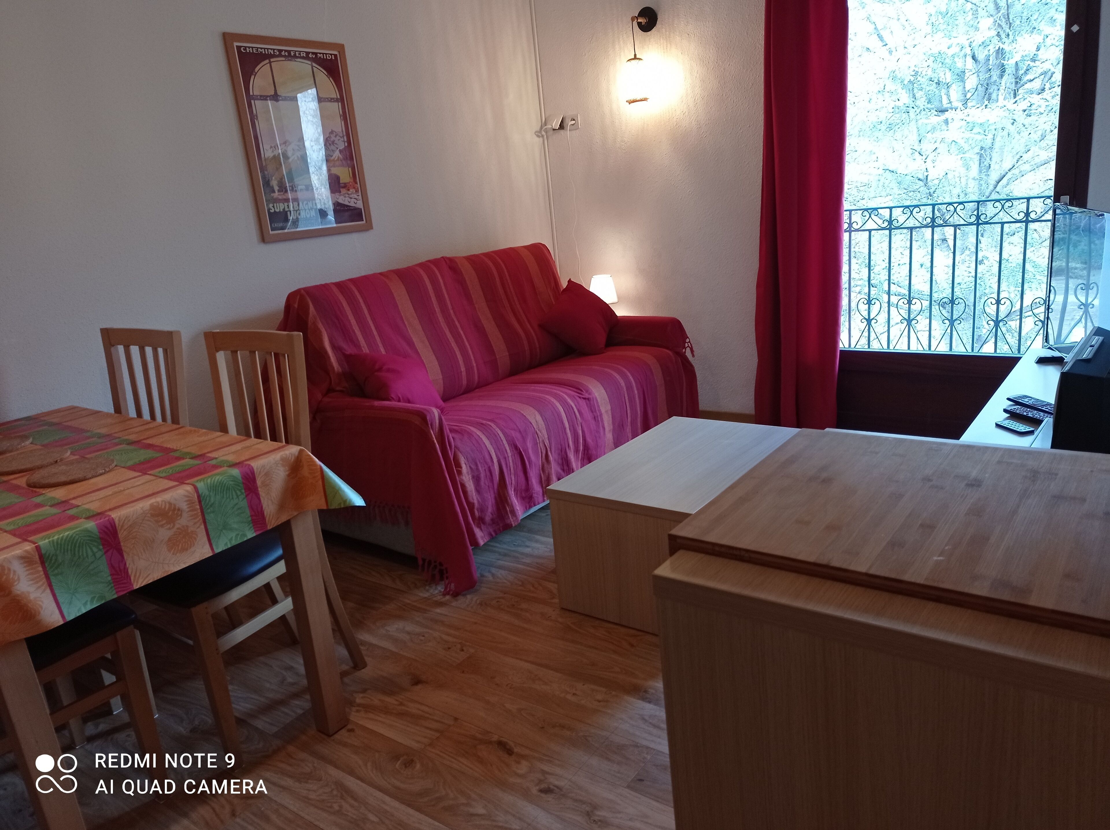 A cozy living room with a red sofa, wooden dining table, and chairs. A small table with a lamp is next to the sofa.