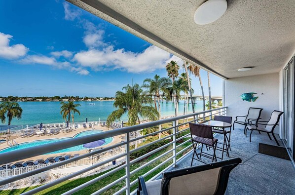 Balcón con vista a una piscina y una playa, equipado con sillas y una mesa.