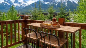 A wooden deck with a table and chairs, a bottle of liquor, and glasses on a tray, with a mountainous landscape in the background. - Lavish Mountain Condo - Amazing Views! (Canmore)