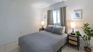 A bedroom with a bed, two bedside tables with lamps, a window with curtains, and a wall-mounted picture. - Fillmore Unit 10 | 2BR/1BA (Cape Canaveral)