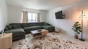 A modern living room with a sectional sofa, a coffee table, a flat-screen TV mounted on the wall, and a floor lamp. - Fillmore Unit 10 | 2BR/1BA (Cape Canaveral)