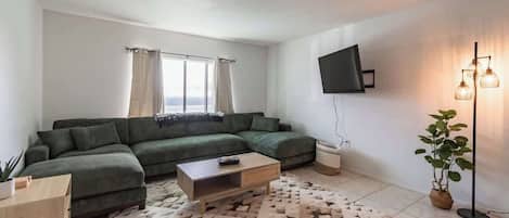 A modern living room with a sectional sofa, a coffee table, a flat-screen TV mounted on the wall, and a floor lamp.