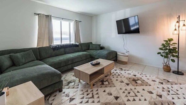 A living room with a green sectional sofa, a wooden coffee table, a flat-screen TV mounted on the wall, a patterned rug, and a potted plant. - Fillmore Unit 10 | 2BR/1BA (Cape Canaveral)