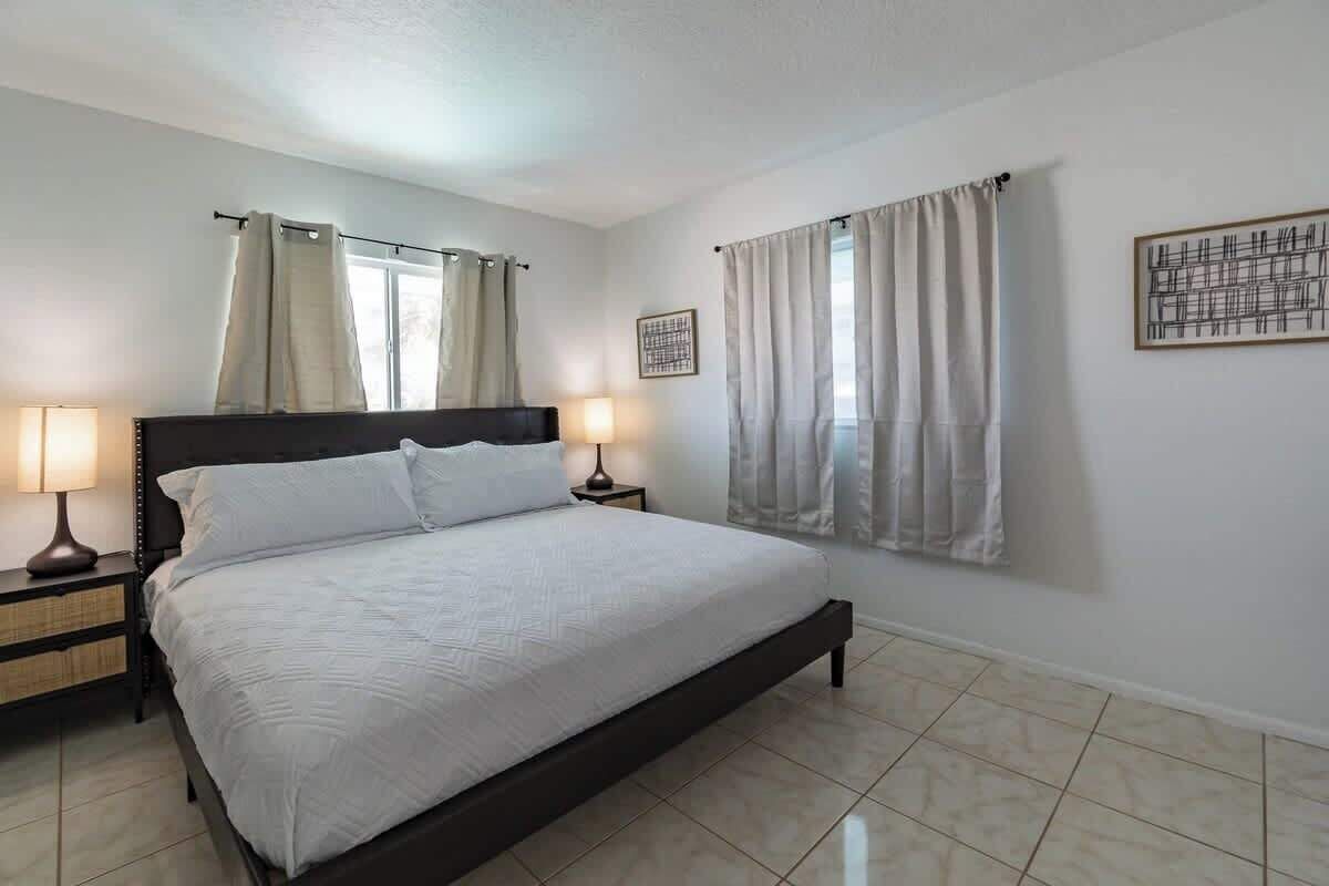 A bedroom with a bed, two bedside tables, a nightstand, a window with curtains, and a framed picture on the wall.