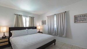 A bedroom with a bed, two bedside tables, a nightstand, a window with curtains, and a framed picture on the wall. - Fillmore Unit 10 | 2BR/1BA (Cape Canaveral)