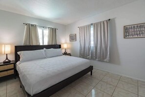 A bedroom with a bed, two bedside tables, a nightstand, a window with curtains, and a framed picture on the wall. - Fillmore Unit 10 | 2BR/1BA (Cape Canaveral)