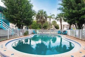 En swimmingpool med en kant som är klädd med kakel, omgiven av ett vitt staket, och träd i bakgrunden.