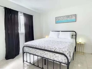 A single bed with a metal frame, a white and gray patterned bedspread, black curtains, a wall-mounted light, and a framed picture above the bed. - Fillmore Unit #1 | 2BR/1BA (Cape Canaveral)