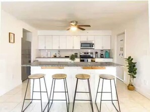 A modern kitchen with white cabinets, a central island with stools, and a ceiling fan. - Fillmore Unit #1 | 2BR/1BA (Cape Canaveral)