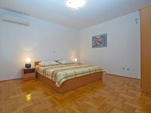 A bedroom with a wooden floor, a bed with a striped bedspread, two bedside lamps, a nightstand, an air conditioning unit, and a wall-mounted painting. - Holiday apartment with air conditioning (Pula)