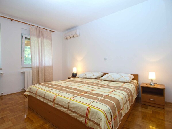 A bedroom with a bed, two bedside tables, a window with curtains, an air conditioning unit, and a radiator. - Holiday apartment with air conditioning (Pula)