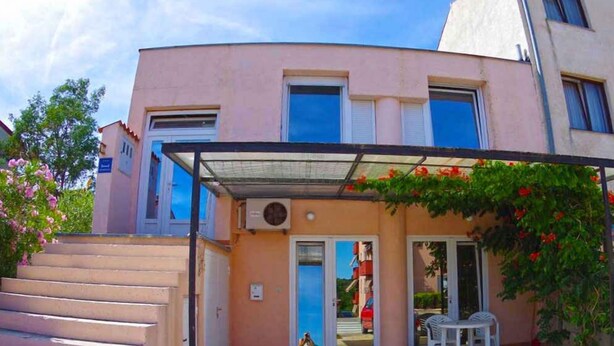 A building with a pink facade, a covered entrance with a glass door, and a small patio area with two chairs.